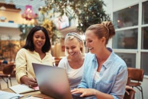 Group of diverse businesswomen smiling while working on a laptop together at a table in an office complex lounge